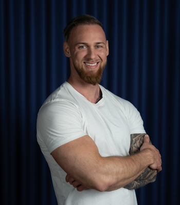 Anders Koch, manuel behandler hos Black Swan Lifestyle. Anders står i en hvid t-shirt med armene over kors og smiler foran en baggrund af et mørkeblåt gardin